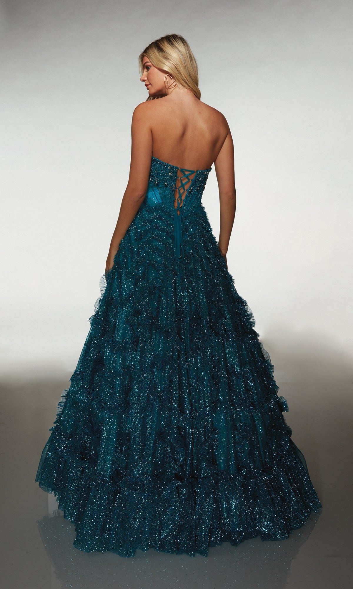 A woman faces away, wearing the Alyce 62132 long formal dress with a strapless lace-up back and a glitter tulle skirt in ruffled, floor-length layers.